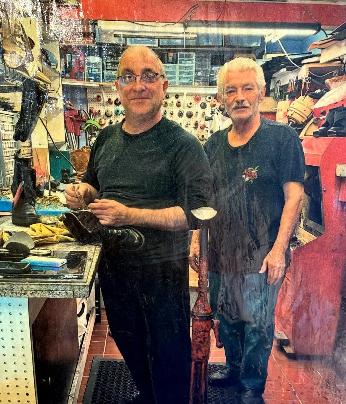 Two of the team at the workbench inside Robert's Shoe Repair, one holding a boot in progress, with lasts, tools and finished shoes around them